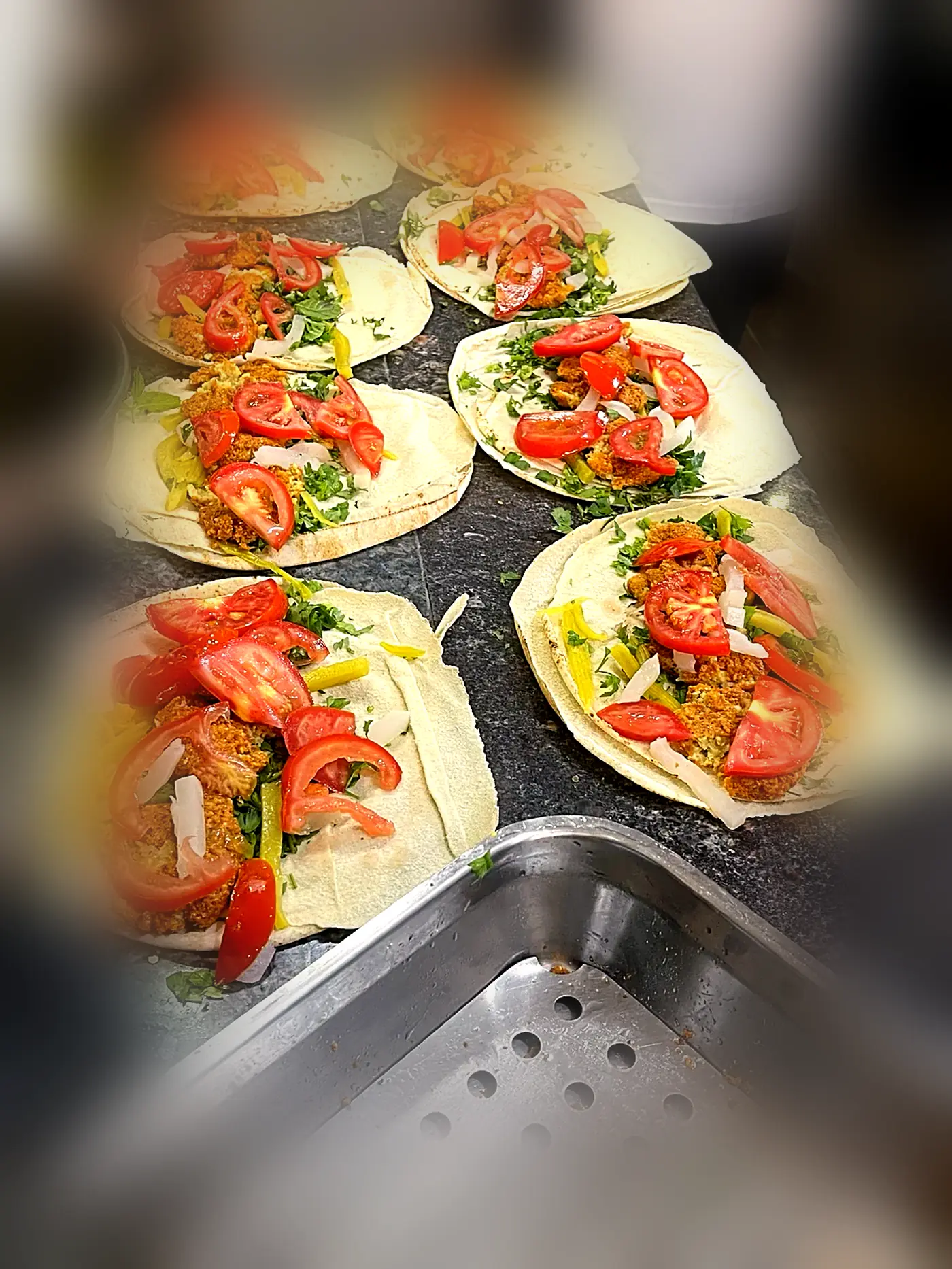 A line of open flatbreads being filled with falafel, tomato, parsley and pickles